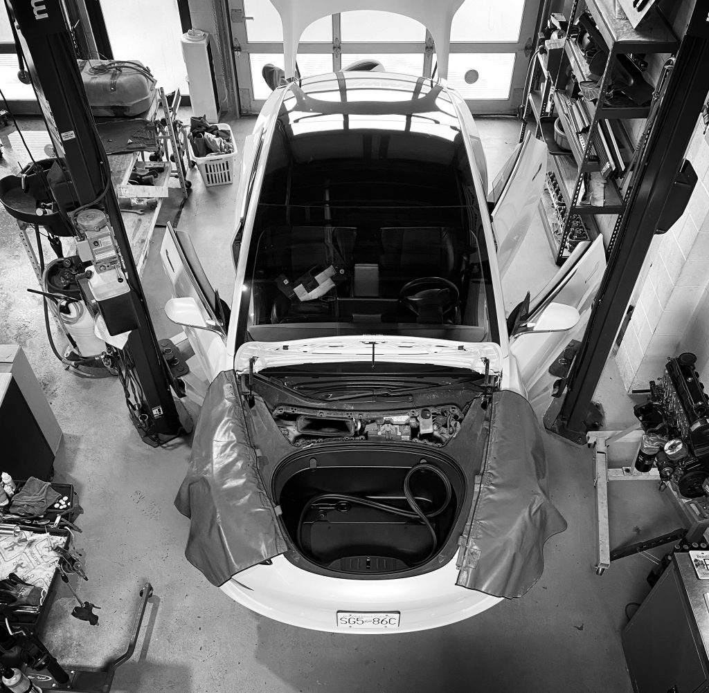 Bird's eye image of a Tesla Model 3 on a hoist in a work shop with the hood open. 
