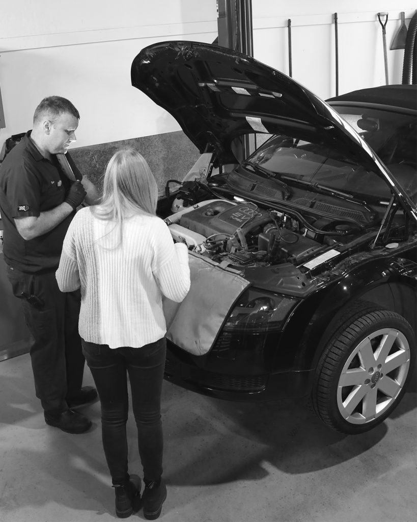 Technician and Service Advisor in front of an Audi with the hood open discussing work on the vehicle in a work shop. 