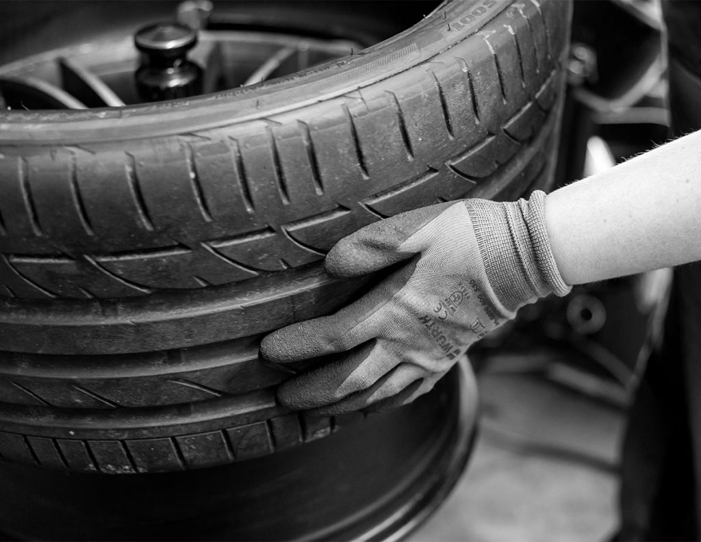 close up black and white image of a technician, wearing gloves, handling a vehicle tire. 